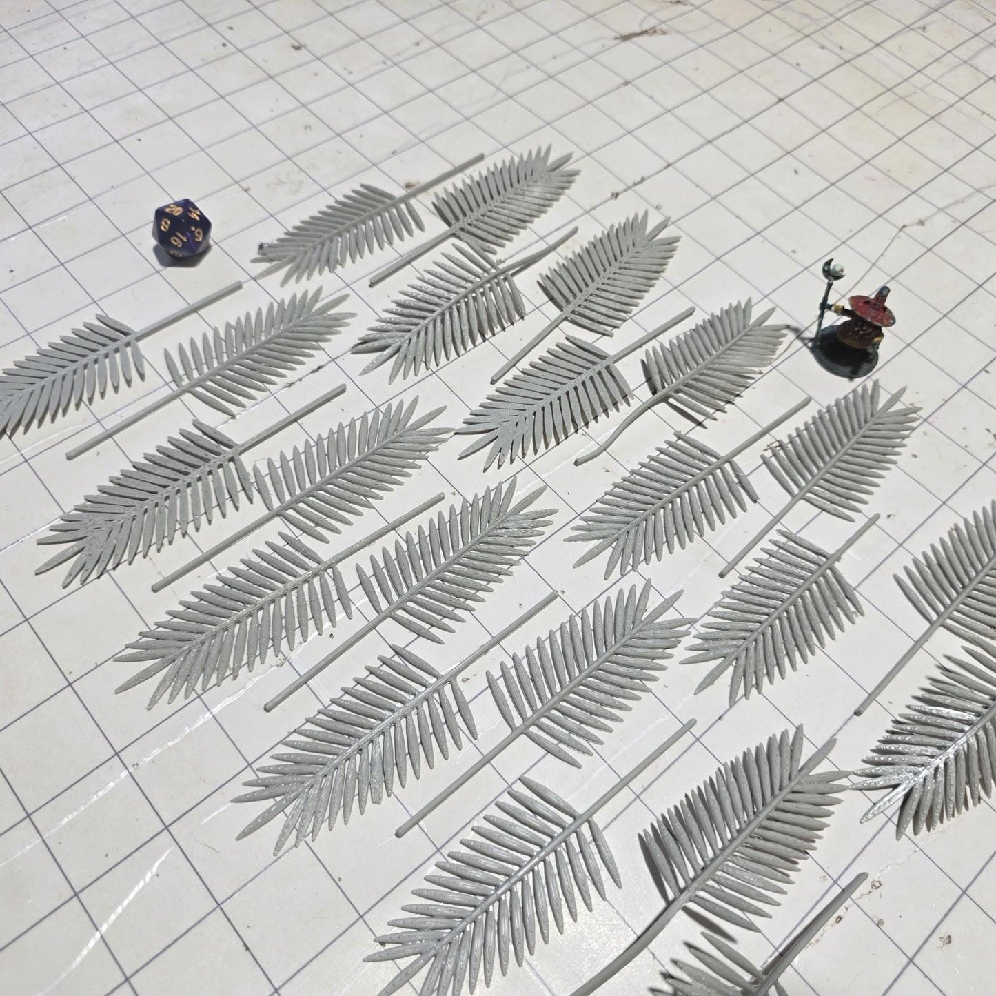 Laid across a 1" grid, these 28mm-scale palm fronds form a tropical spread waiting to be shaped and painted. With identical sprigs that bend under heat, they’re perfect for scratch-building palm trees, decorating temple ruins, or fleshing out dense jungle scenes. A miniature wizard and dice provide a sense of scale, highlighting their table-ready size.