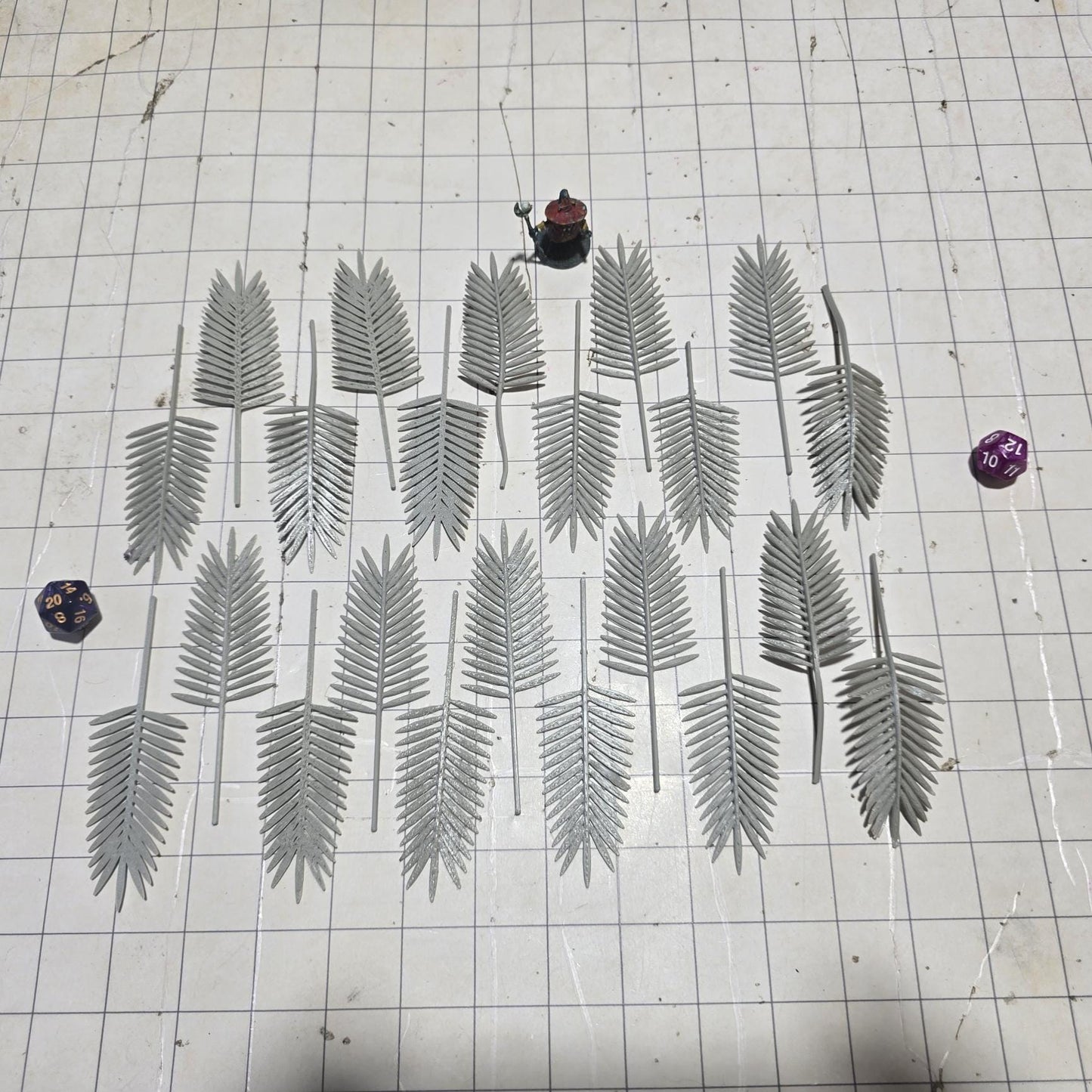 Laid across a 1" grid, these 28mm-scale palm fronds form a tropical spread waiting to be shaped and painted. With identical sprigs that bend under heat, they’re perfect for scratch-building palm trees, decorating temple ruins, or fleshing out dense jungle scenes. A miniature wizard and dice provide a sense of scale, highlighting their table-ready size.