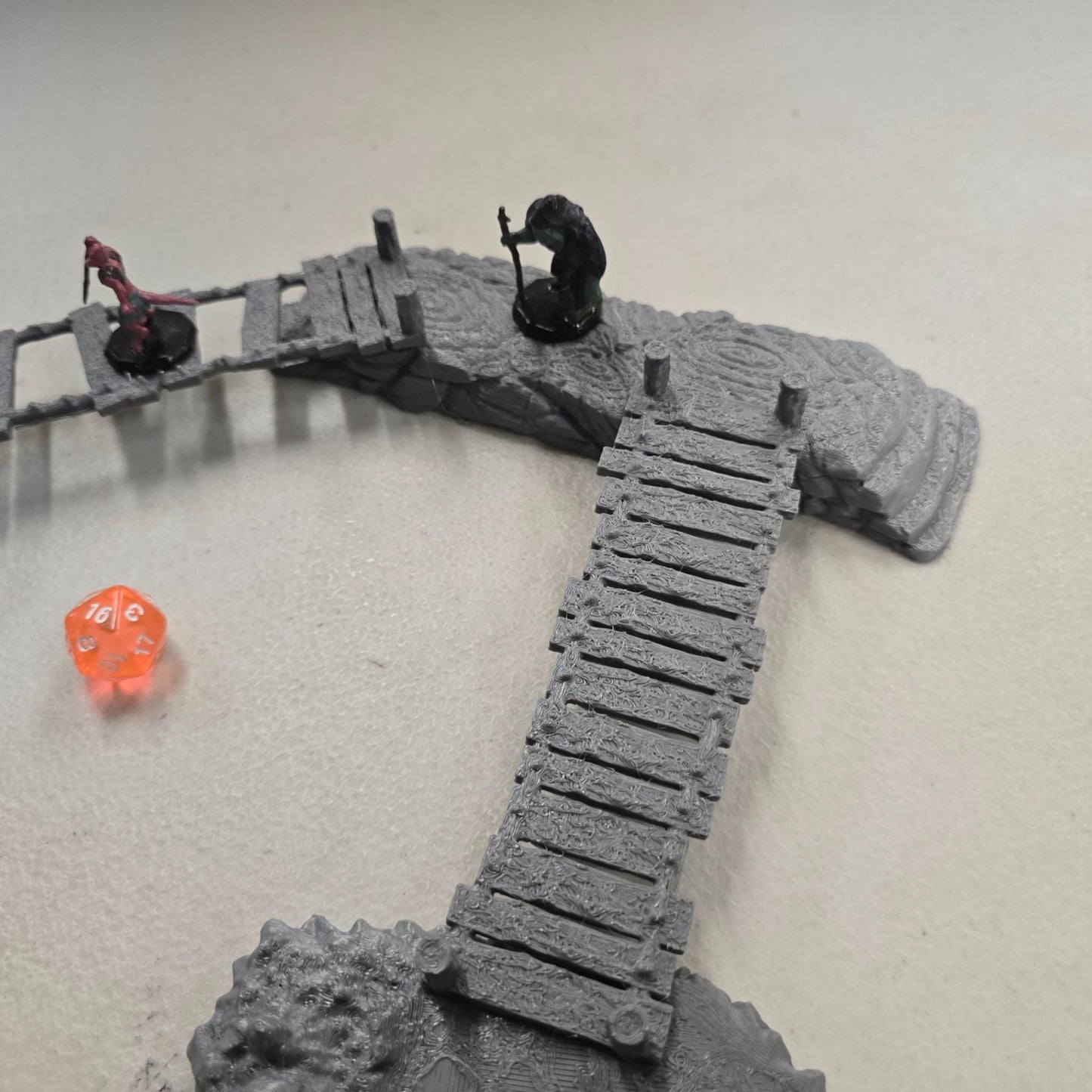 cavern terrain scene made from unpainted gray PLA shows jagged stone platforms connected by narrow wooden plank bridges. Stalagmites rise from the rugged cave floor while a carved stone landing platform forms one end of the crossing. The terrain is scaled for 28 mm tabletop terrain and designed to align with a 1-inch grid for DnD and wargaming encounters. The curved bridge route between rocky platforms creates a dramatic underground battlefield filled with narrow paths and ambush points.