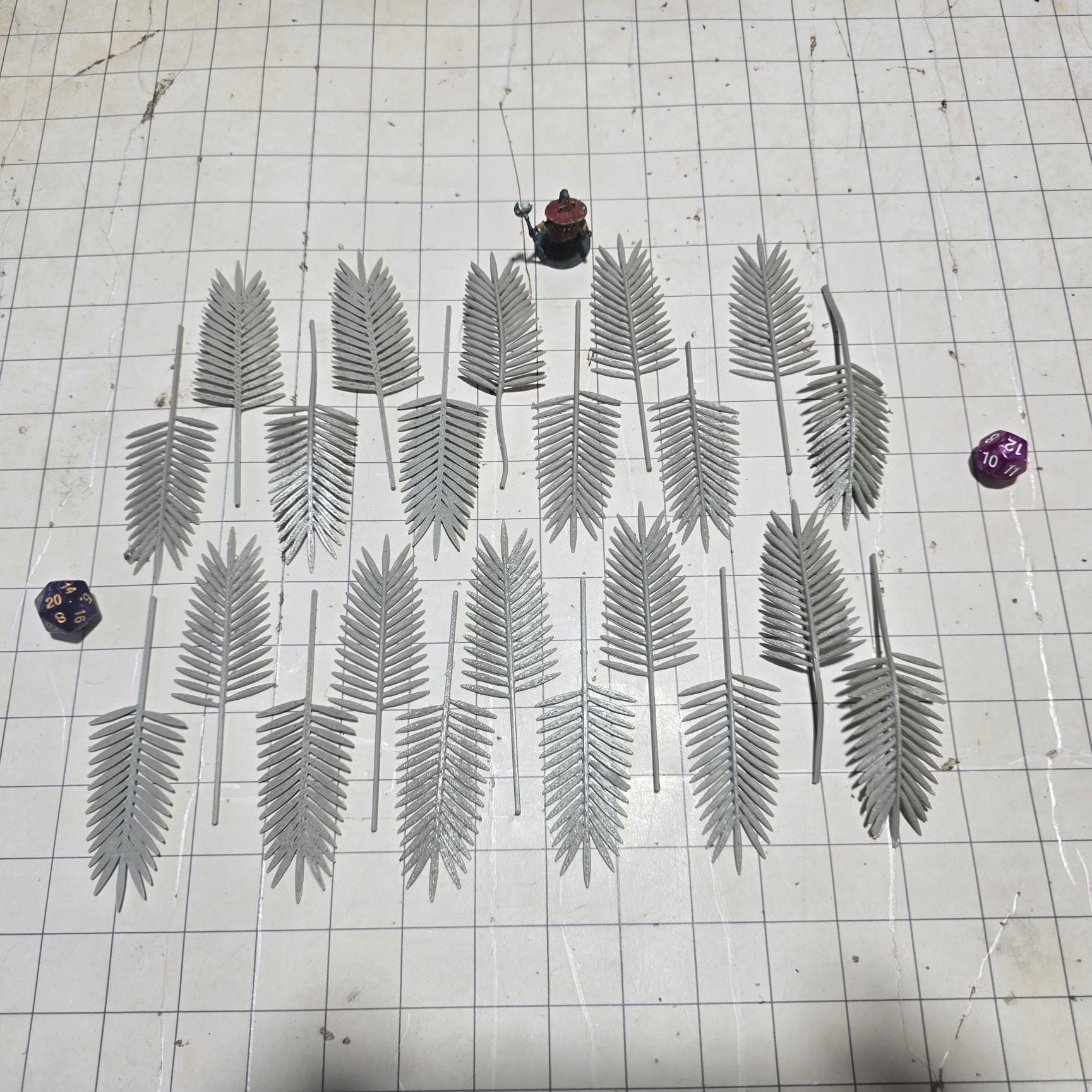 Laid across a 1" grid, these 28mm-scale palm fronds form a tropical spread waiting to be shaped and painted. With identical sprigs that bend under heat, they’re perfect for scratch-building palm trees, decorating temple ruins, or fleshing out dense jungle scenes. A miniature wizard and dice provide a sense of scale, highlighting their table-ready size.