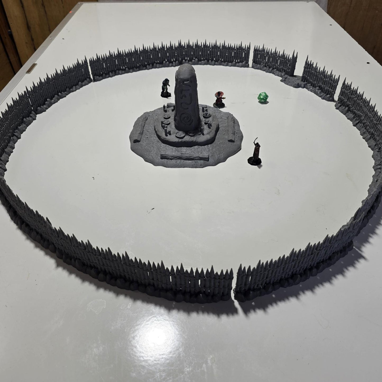 Unpainted gray PLA captures the rough ground, carved runestone, and jagged timber spikes of this 28 mm tabletop terrain set. The monolith rises from a layered base scattered with simple offerings, while the spiked walls form a full circle around the site. Each piece shows the subtle lines of 3D printed construction, ready for primer. Designed for DnD / Pathfinder / wargaming on a 1-inch grid, it stands as a stark landmark on any table.