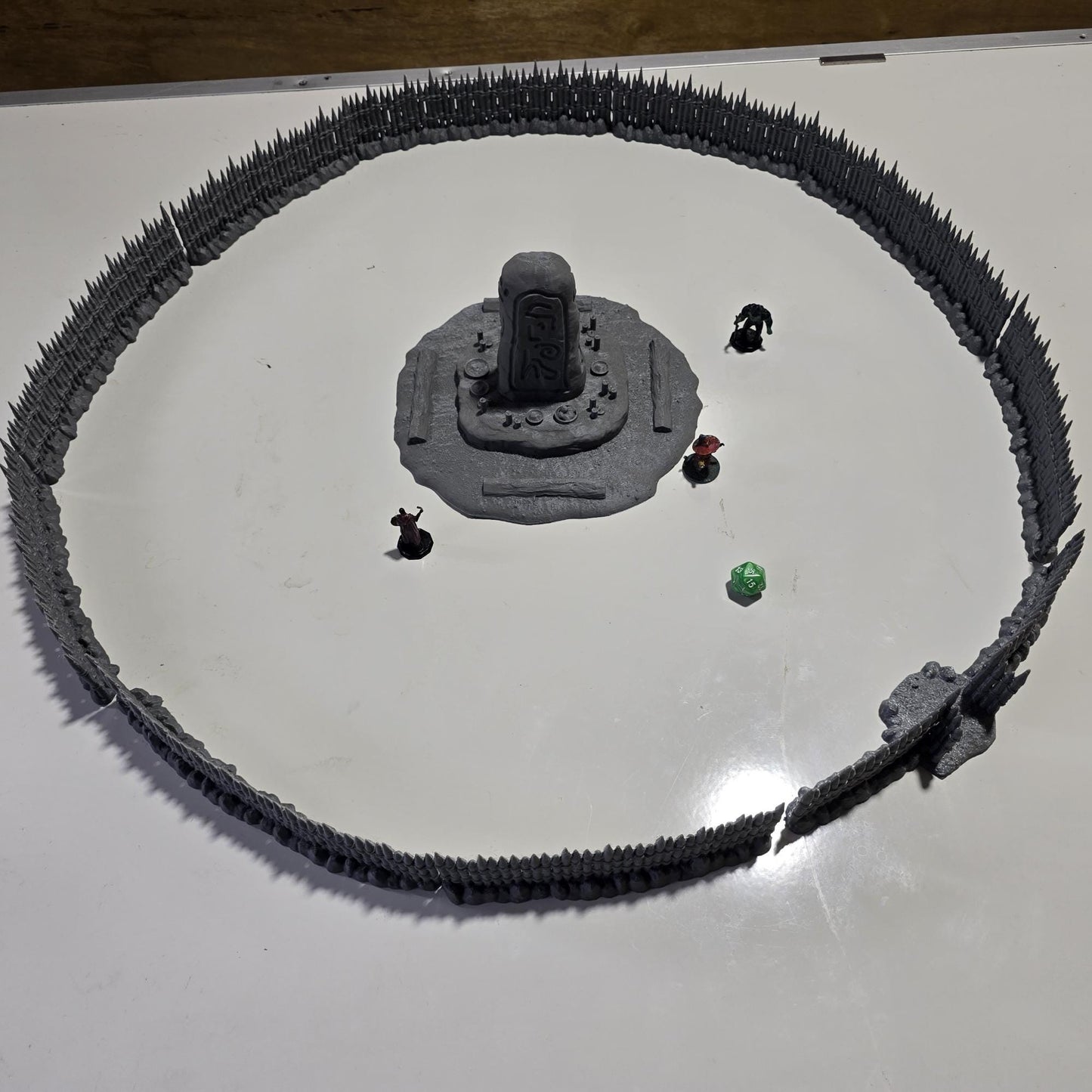 Unpainted gray PLA captures the rough ground, carved runestone, and jagged timber spikes of this 28 mm tabletop terrain set. The monolith rises from a layered base scattered with simple offerings, while the spiked walls form a full circle around the site. Each piece shows the subtle lines of 3D printed construction, ready for primer. Designed for DnD / Pathfinder / wargaming on a 1-inch grid, it stands as a stark landmark on any table.