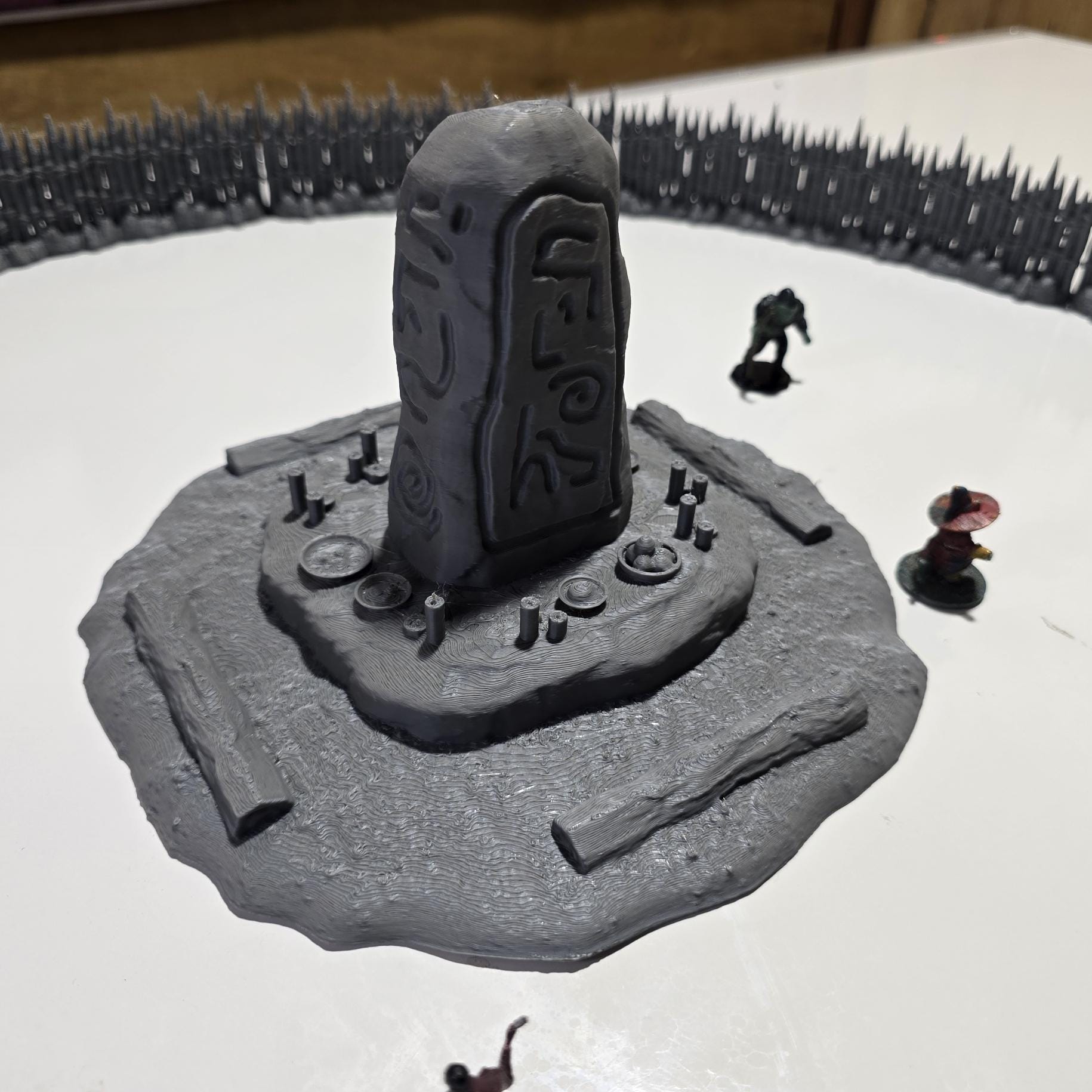 Unpainted gray PLA captures the rough ground, carved runestone, and jagged timber spikes of this 28 mm tabletop terrain set. The monolith rises from a layered base scattered with simple offerings, while the spiked walls form a full circle around the site. Each piece shows the subtle lines of 3D printed construction, ready for primer. Designed for DnD / Pathfinder / wargaming on a 1-inch grid, it stands as a stark landmark on any table.