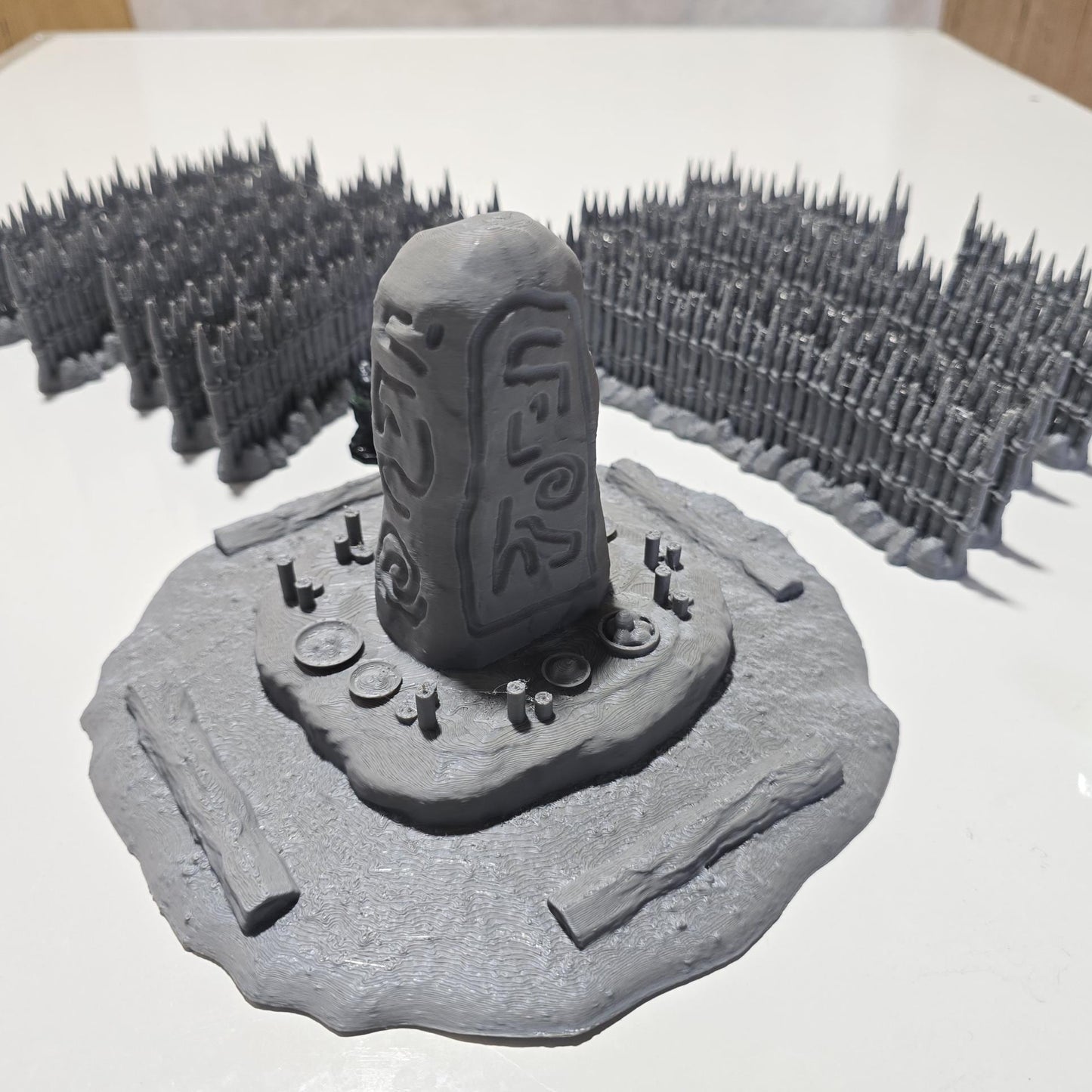 Unpainted gray PLA captures the rough ground, carved runestone, and jagged timber spikes of this 28 mm tabletop terrain set. The monolith rises from a layered base scattered with simple offerings, while the spiked walls form a full circle around the site. Each piece shows the subtle lines of 3D printed construction, ready for primer. Designed for DnD / Pathfinder / wargaming on a 1-inch grid, it stands as a stark landmark on any table.