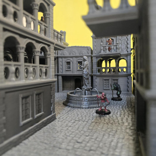 A sprawling 3D printed fantasy city rises across the table, assembled from unpainted gray PLA buildings arranged along cobblestone streets and balcony-lined structures. Arched windows, terrace railings, and stacked stone walls create layered architecture scaled for 28 mm tabletop terrain and a 1-inch grid. In the center plaza, a sculpted fountain statue anchors the streets while rooftops and balconies overlook narrow alleys and open squares.