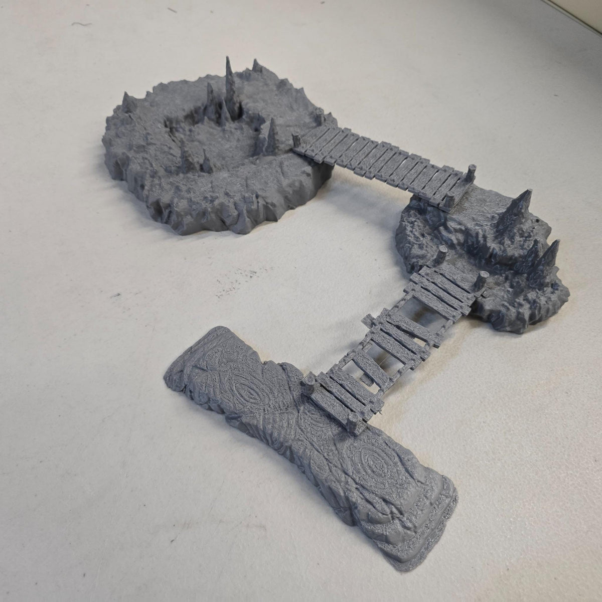 Cavern terrain scene made from unpainted gray PLA shows jagged cave platforms connected by narrow wooden bridges. Stalagmites rise from the rocky cavern floor while a carved stone slab landing forms one end of the crossing. The terrain is scaled for 28 mm tabletop terrain and designed to align with a 1-inch grid for DnD, Pathfinder, and wargaming encounters. The suspended bridges and layered cave rock create a dramatic underground battlefield filled with narrow paths and ambush points.