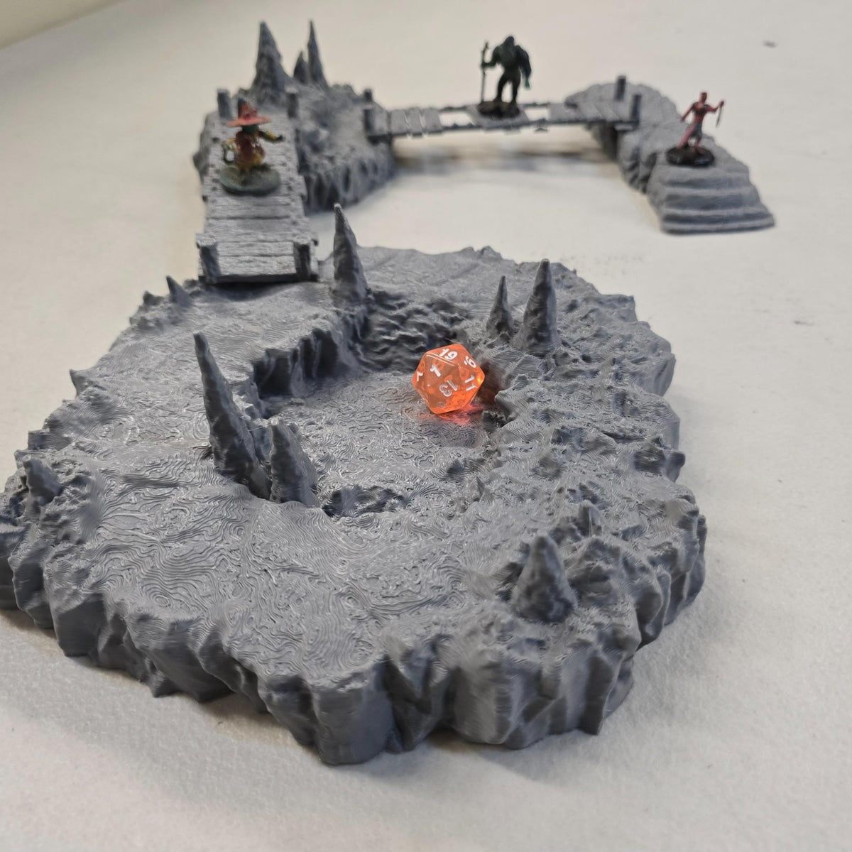 Cavern terrain scene made from unpainted gray PLA shows jagged cave platforms connected by narrow wooden bridges. Stalagmites rise from the rocky cavern floor while a carved stone slab landing forms one end of the crossing. The terrain is scaled for 28 mm tabletop terrain and designed to align with a 1-inch grid for DnD, Pathfinder, and wargaming encounters. The suspended bridges and layered cave rock create a dramatic underground battlefield filled with narrow paths and ambush points.