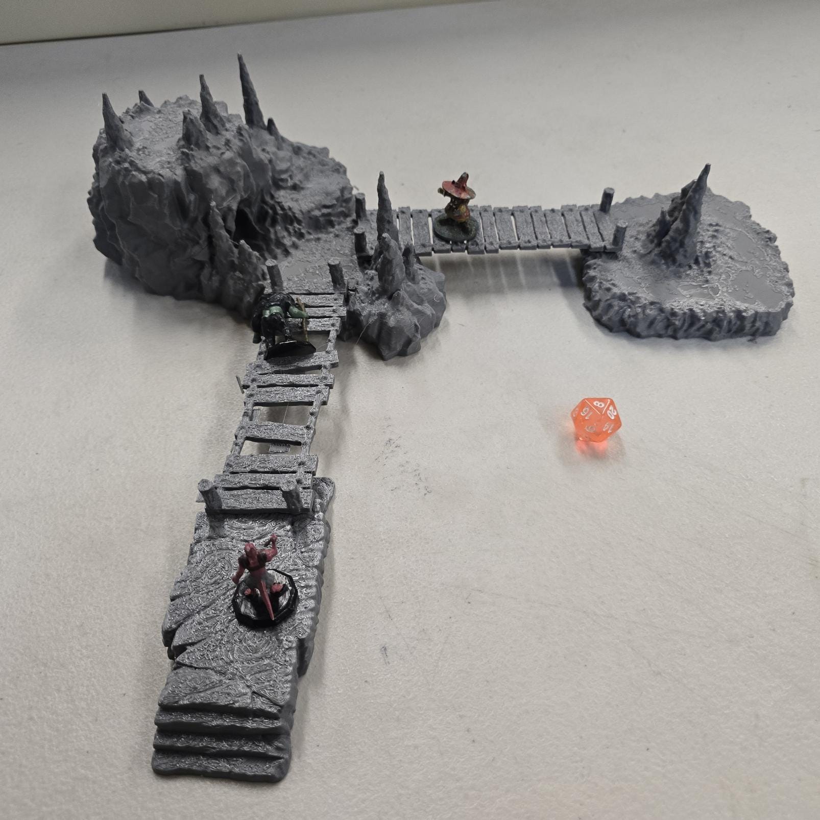 Cavern terrain encounter made from unpainted gray PLA shows a rocky cave entrance surrounded by tall stalagmites and connected by narrow wooden plank bridges. The scene includes a raised stone landing platform and rugged cavern platforms forming a dangerous path across the underground chamber. Scaled for 28 mm tabletop terrain and aligned to a 1-inch grid. The suspended bridges and jagged cavern stone create a tense underground crossing perfect for ambushes and guarded dungeon passages.