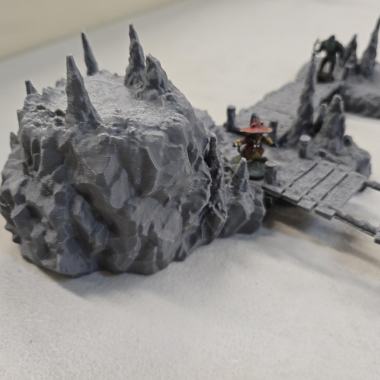 Cavern terrain encounter made from unpainted gray PLA shows a rocky cave entrance surrounded by tall stalagmites and connected by narrow wooden plank bridges. The scene includes a raised stone landing platform and rugged cavern platforms forming a dangerous path across the underground chamber. Scaled for 28 mm tabletop terrain and aligned to a 1-inch grid. The suspended bridges and jagged cavern stone create a tense underground crossing perfect for ambushes and guarded dungeon passages.