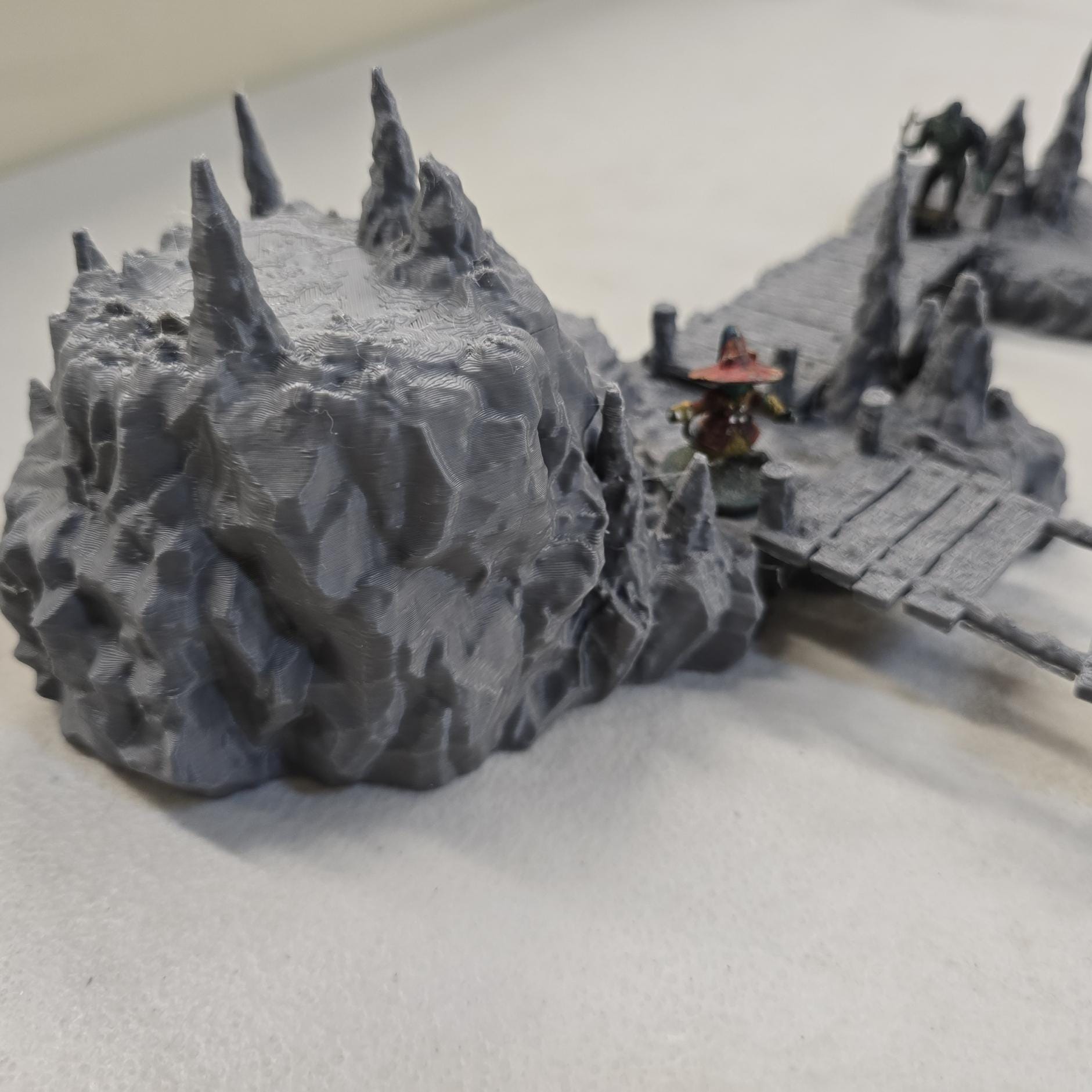 Cavern terrain encounter made from unpainted gray PLA shows a rocky cave entrance surrounded by tall stalagmites and connected by narrow wooden plank bridges. The scene includes a raised stone landing platform and rugged cavern platforms forming a dangerous path across the underground chamber. Scaled for 28 mm tabletop terrain and aligned to a 1-inch grid. The suspended bridges and jagged cavern stone create a tense underground crossing perfect for ambushes and guarded dungeon passages.