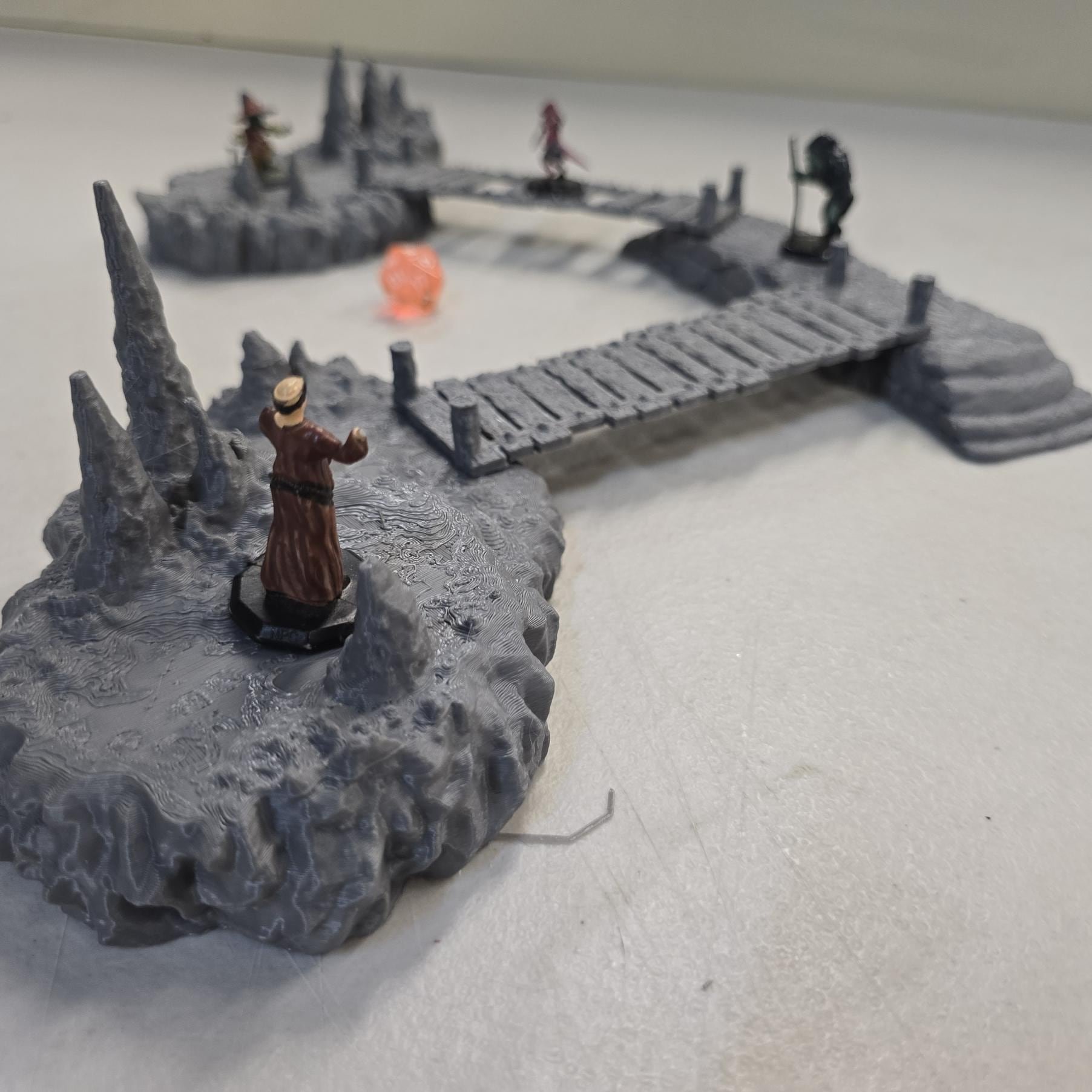 cavern terrain scene made from unpainted gray PLA shows jagged stone platforms connected by narrow wooden plank bridges. Stalagmites rise from the rugged cave floor while a carved stone landing platform forms one end of the crossing. The terrain is scaled for 28 mm tabletop terrain and designed to align with a 1-inch grid for DnD and wargaming encounters. The curved bridge route between rocky platforms creates a dramatic underground battlefield filled with narrow paths and ambush points.