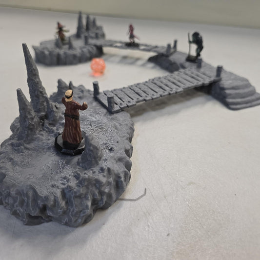 cavern terrain scene made from unpainted gray PLA shows jagged stone platforms connected by narrow wooden plank bridges. Stalagmites rise from the rugged cave floor while a carved stone landing platform forms one end of the crossing. The terrain is scaled for 28 mm tabletop terrain and designed to align with a 1-inch grid for DnD and wargaming encounters. The curved bridge route between rocky platforms creates a dramatic underground battlefield filled with narrow paths and ambush points.