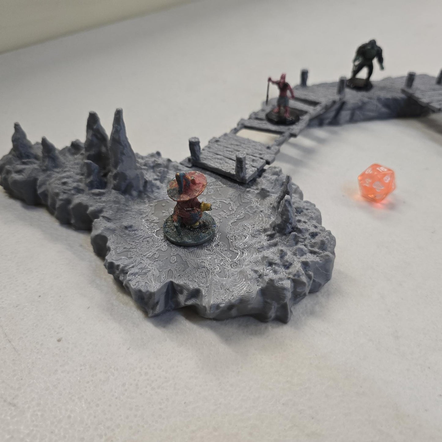 cavern terrain scene made from unpainted gray PLA shows jagged stone platforms connected by narrow wooden plank bridges. Stalagmites rise from the rugged cave floor while a carved stone landing platform forms one end of the crossing. The terrain is scaled for 28 mm tabletop terrain and designed to align with a 1-inch grid for DnD and wargaming encounters. The curved bridge route between rocky platforms creates a dramatic underground battlefield filled with narrow paths and ambush points.