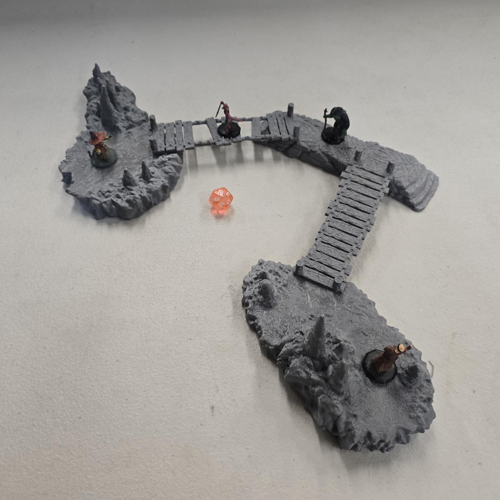 cavern terrain scene made from unpainted gray PLA shows jagged stone platforms connected by narrow wooden plank bridges. Stalagmites rise from the rugged cave floor while a carved stone landing platform forms one end of the crossing. The terrain is scaled for 28 mm tabletop terrain and designed to align with a 1-inch grid for DnD and wargaming encounters. The curved bridge route between rocky platforms creates a dramatic underground battlefield filled with narrow paths and ambush points.