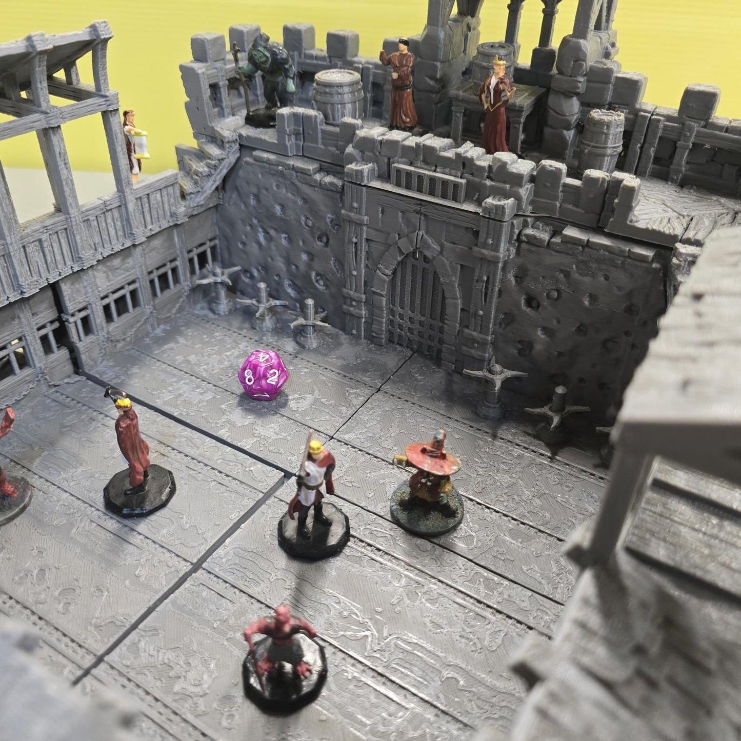 Thick stone walls, iron-barred gates, arched doorways, and elevated wooden galleries surround a detailed courtyard floor divided into grid-aligned tiles. Removable roof sections reveal interior balconies and bolt-scarred arena walls designed for tactical movement and line-of-sight control. The enclosed design creates a brutal city combat zone ready for close-quarters encounters.