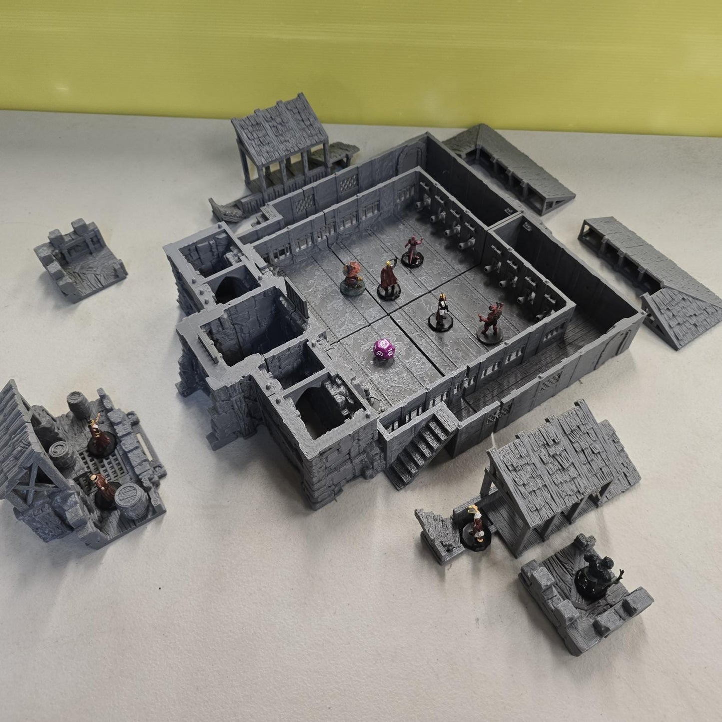 Thick stone walls, iron-barred gates, arched doorways, and elevated wooden galleries surround a detailed courtyard floor divided into grid-aligned tiles. Removable roof sections reveal interior balconies and bolt-scarred arena walls designed for tactical movement and line-of-sight control. The enclosed design creates a brutal city combat zone ready for close-quarters encounters.