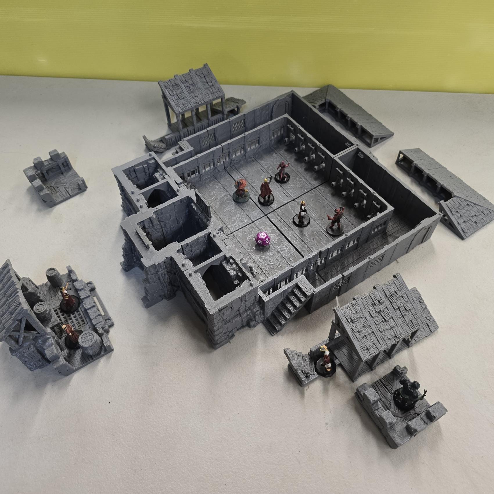 Thick stone walls, iron-barred gates, arched doorways, and elevated wooden galleries surround a detailed courtyard floor divided into grid-aligned tiles. Removable roof sections reveal interior balconies and bolt-scarred arena walls designed for tactical movement and line-of-sight control. The enclosed design creates a brutal city combat zone ready for close-quarters encounters.