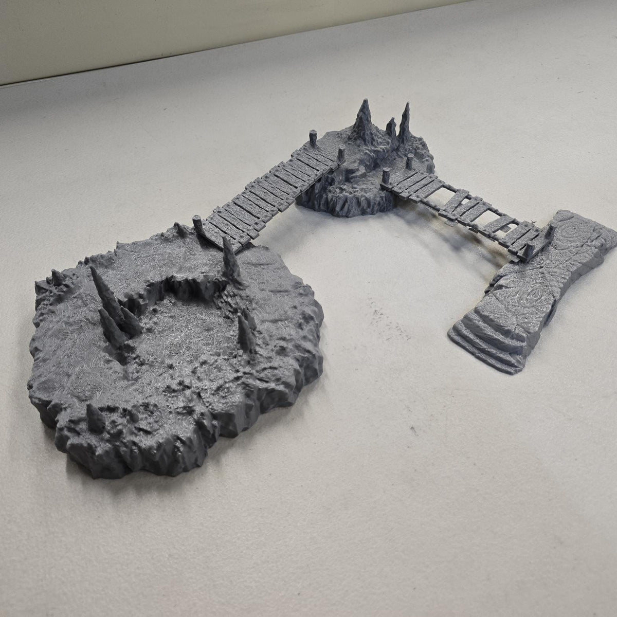 Cavern terrain scene made from unpainted gray PLA shows jagged cave platforms connected by narrow wooden bridges. Stalagmites rise from the rocky cavern floor while a carved stone slab landing forms one end of the crossing. The terrain is scaled for 28 mm tabletop terrain and designed to align with a 1-inch grid for DnD, Pathfinder, and wargaming encounters. The suspended bridges and layered cave rock create a dramatic underground battlefield filled with narrow paths and ambush points.
