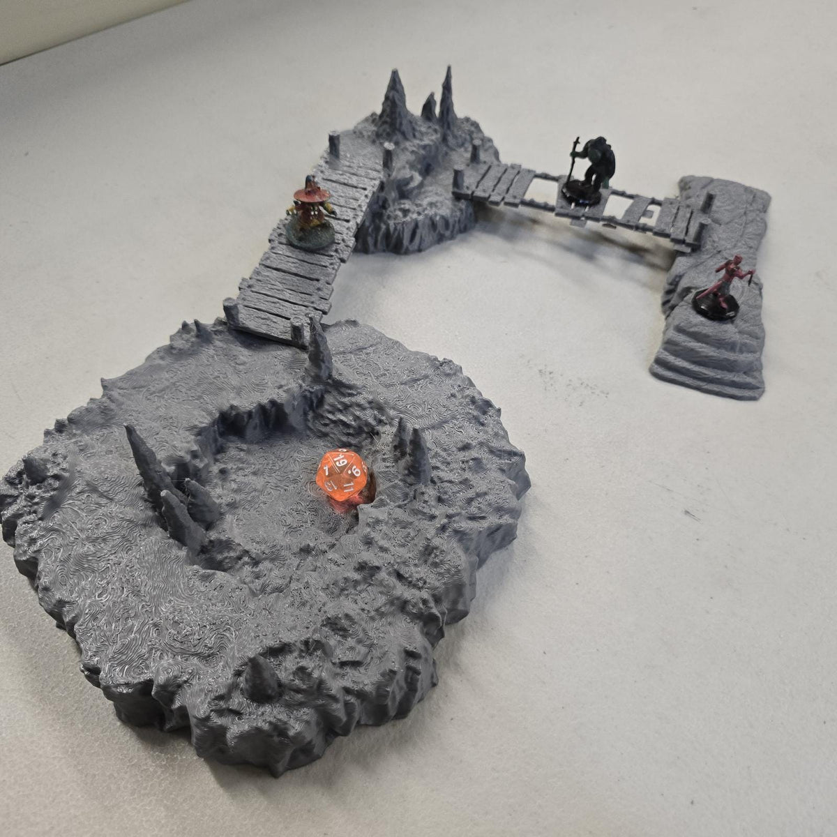 Cavern terrain scene made from unpainted gray PLA shows jagged cave platforms connected by narrow wooden bridges. Stalagmites rise from the rocky cavern floor while a carved stone slab landing forms one end of the crossing. The terrain is scaled for 28 mm tabletop terrain and designed to align with a 1-inch grid for DnD, Pathfinder, and wargaming encounters. The suspended bridges and layered cave rock create a dramatic underground battlefield filled with narrow paths and ambush points.