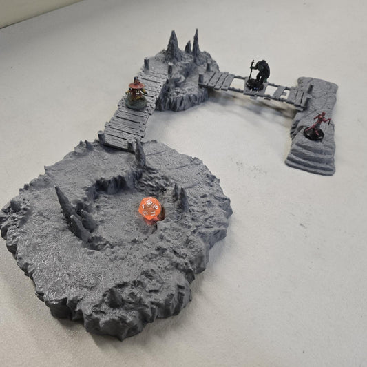 Cavern terrain scene made from unpainted gray PLA shows jagged cave platforms connected by narrow wooden bridges. Stalagmites rise from the rocky cavern floor while a carved stone slab landing forms one end of the crossing. The terrain is scaled for 28 mm tabletop terrain and designed to align with a 1-inch grid for DnD, Pathfinder, and wargaming encounters. The suspended bridges and layered cave rock create a dramatic underground battlefield filled with narrow paths and ambush points.