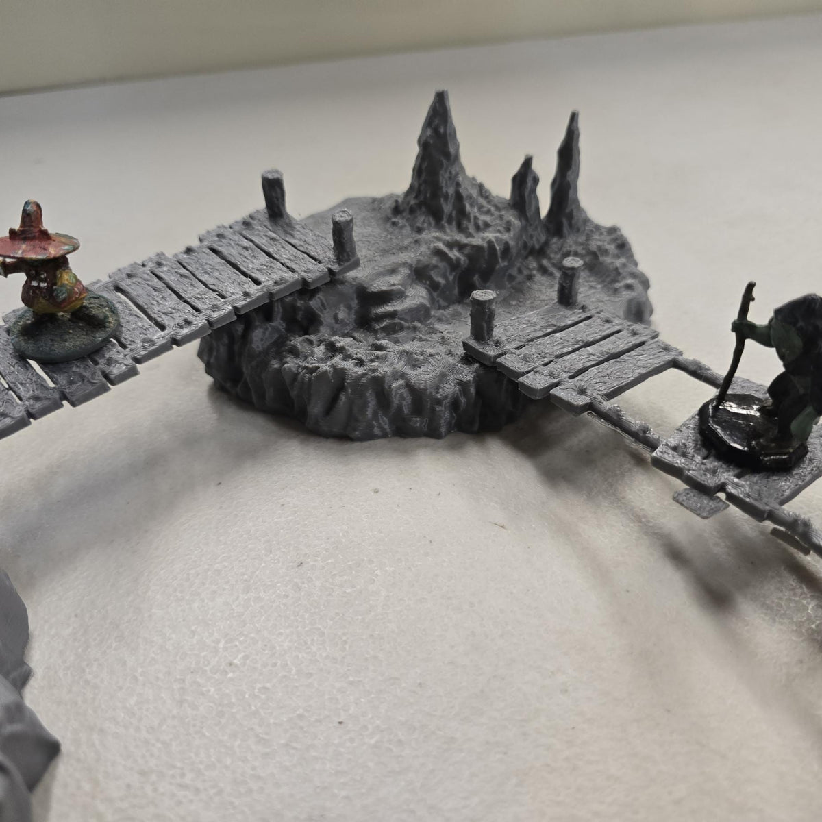 Cavern terrain scene made from unpainted gray PLA shows jagged cave platforms connected by narrow wooden bridges. Stalagmites rise from the rocky cavern floor while a carved stone slab landing forms one end of the crossing. The terrain is scaled for 28 mm tabletop terrain and designed to align with a 1-inch grid for DnD, Pathfinder, and wargaming encounters. The suspended bridges and layered cave rock create a dramatic underground battlefield filled with narrow paths and ambush points.