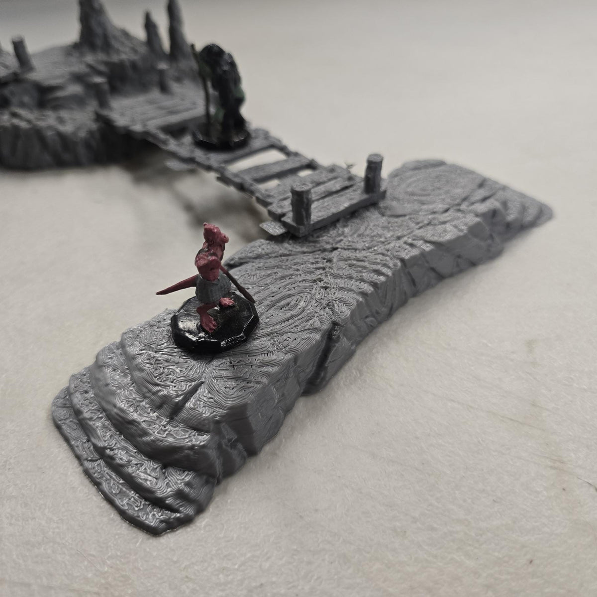 Cavern terrain scene made from unpainted gray PLA shows jagged cave platforms connected by narrow wooden bridges. Stalagmites rise from the rocky cavern floor while a carved stone slab landing forms one end of the crossing. The terrain is scaled for 28 mm tabletop terrain and designed to align with a 1-inch grid for DnD, Pathfinder, and wargaming encounters. The suspended bridges and layered cave rock create a dramatic underground battlefield filled with narrow paths and ambush points.