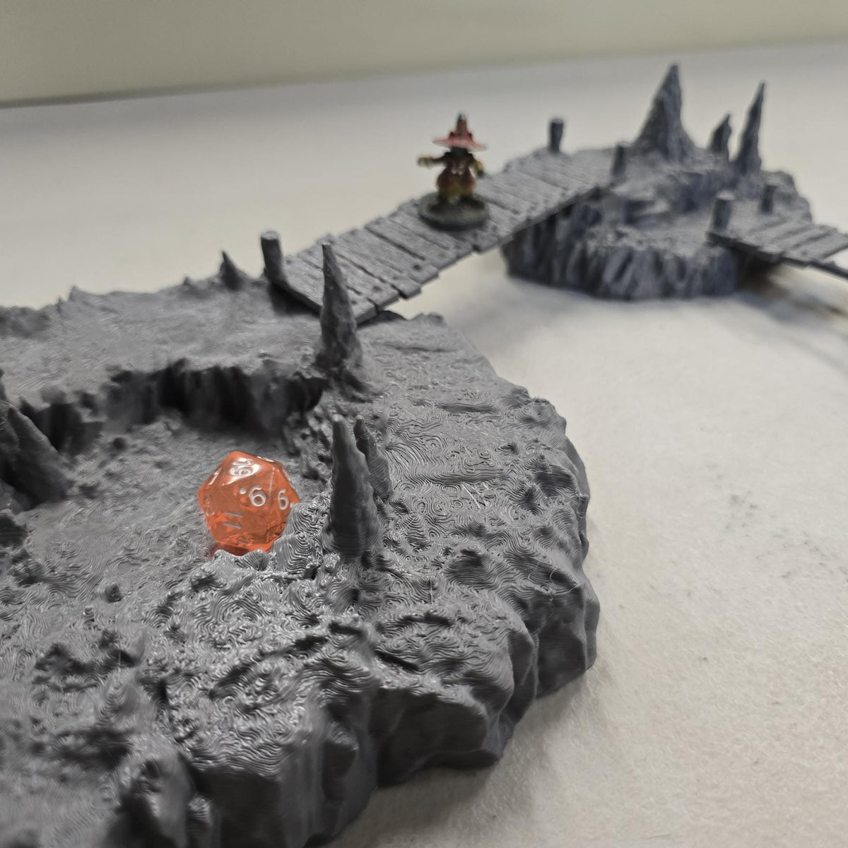 Cavern terrain scene made from unpainted gray PLA shows jagged cave platforms connected by narrow wooden bridges. Stalagmites rise from the rocky cavern floor while a carved stone slab landing forms one end of the crossing. The terrain is scaled for 28 mm tabletop terrain and designed to align with a 1-inch grid for DnD, Pathfinder, and wargaming encounters. The suspended bridges and layered cave rock create a dramatic underground battlefield filled with narrow paths and ambush points.