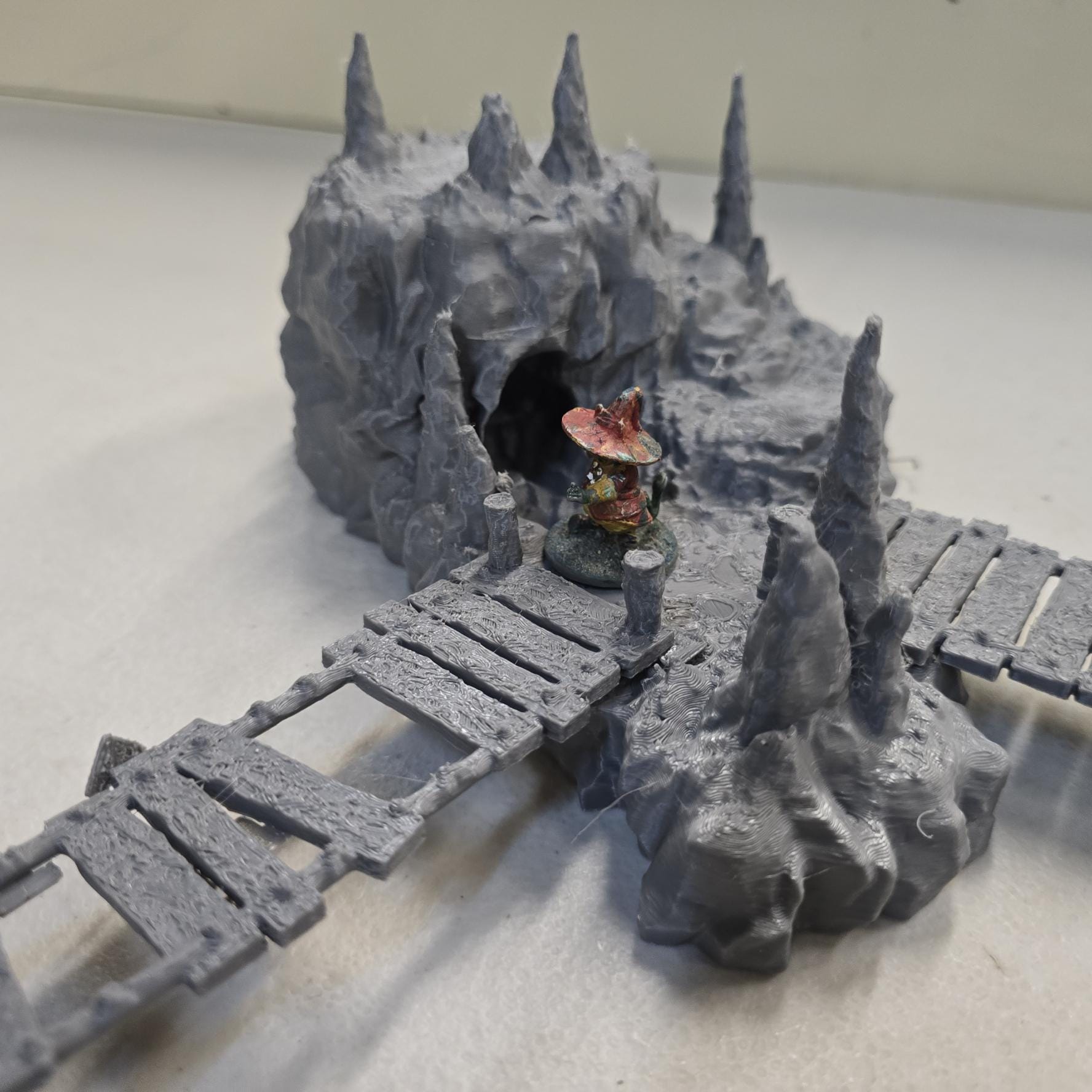 Cavern terrain encounter made from unpainted gray PLA shows a rocky cave entrance surrounded by tall stalagmites and connected by narrow wooden plank bridges. The scene includes a raised stone landing platform and rugged cavern platforms forming a dangerous path across the underground chamber. Scaled for 28 mm tabletop terrain and aligned to a 1-inch grid. The suspended bridges and jagged cavern stone create a tense underground crossing perfect for ambushes and guarded dungeon passages.