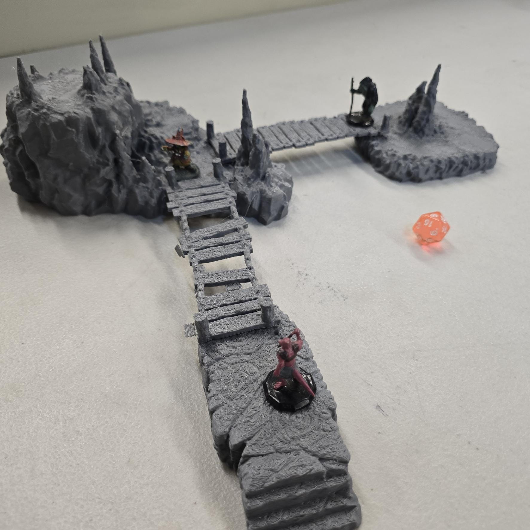 Cavern terrain encounter made from unpainted gray PLA shows a rocky cave entrance surrounded by tall stalagmites and connected by narrow wooden plank bridges. The scene includes a raised stone landing platform and rugged cavern platforms forming a dangerous path across the underground chamber. Scaled for 28 mm tabletop terrain and aligned to a 1-inch grid. The suspended bridges and jagged cavern stone create a tense underground crossing perfect for ambushes and guarded dungeon passages.