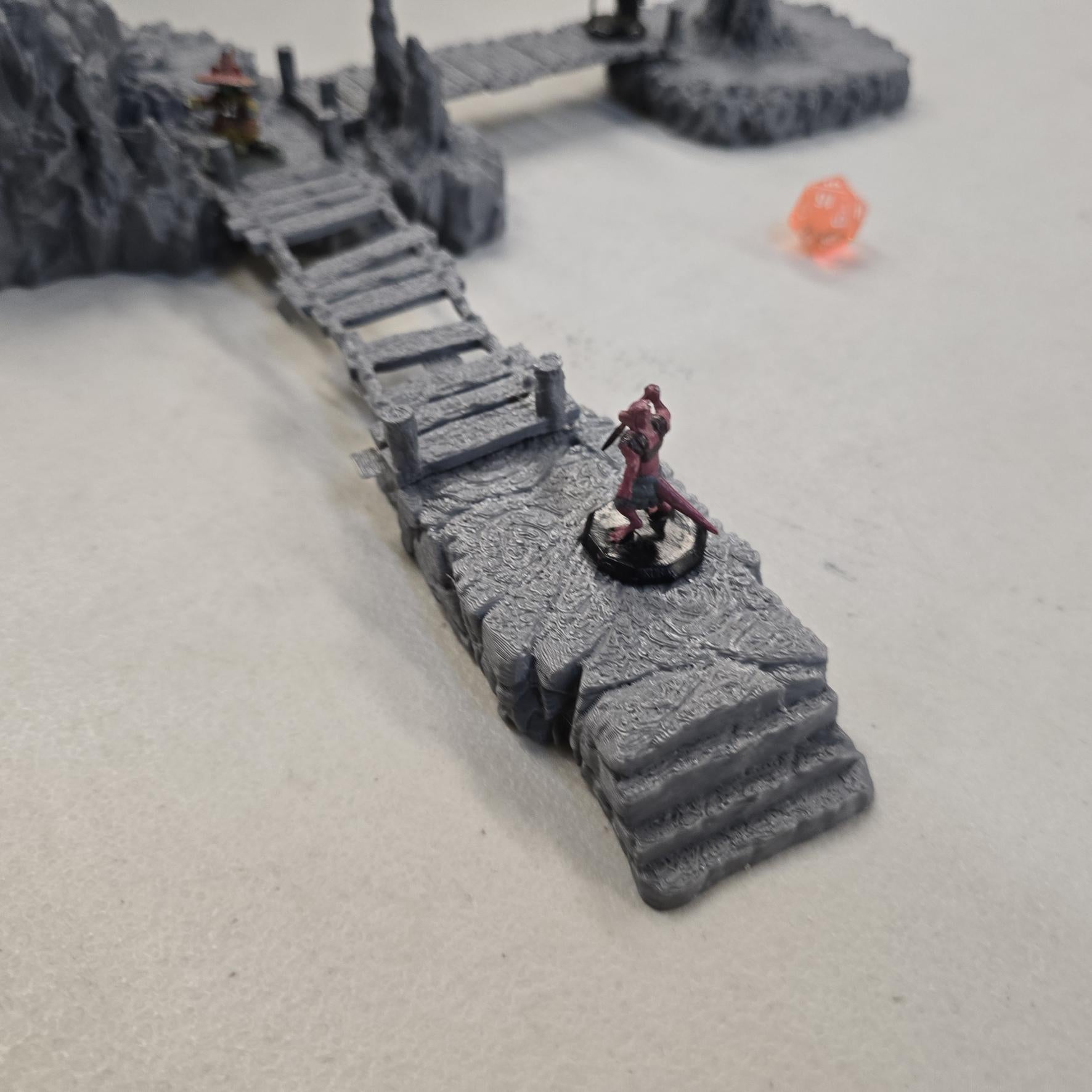 Cavern terrain encounter made from unpainted gray PLA shows a rocky cave entrance surrounded by tall stalagmites and connected by narrow wooden plank bridges. The scene includes a raised stone landing platform and rugged cavern platforms forming a dangerous path across the underground chamber. Scaled for 28 mm tabletop terrain and aligned to a 1-inch grid. The suspended bridges and jagged cavern stone create a tense underground crossing perfect for ambushes and guarded dungeon passages.