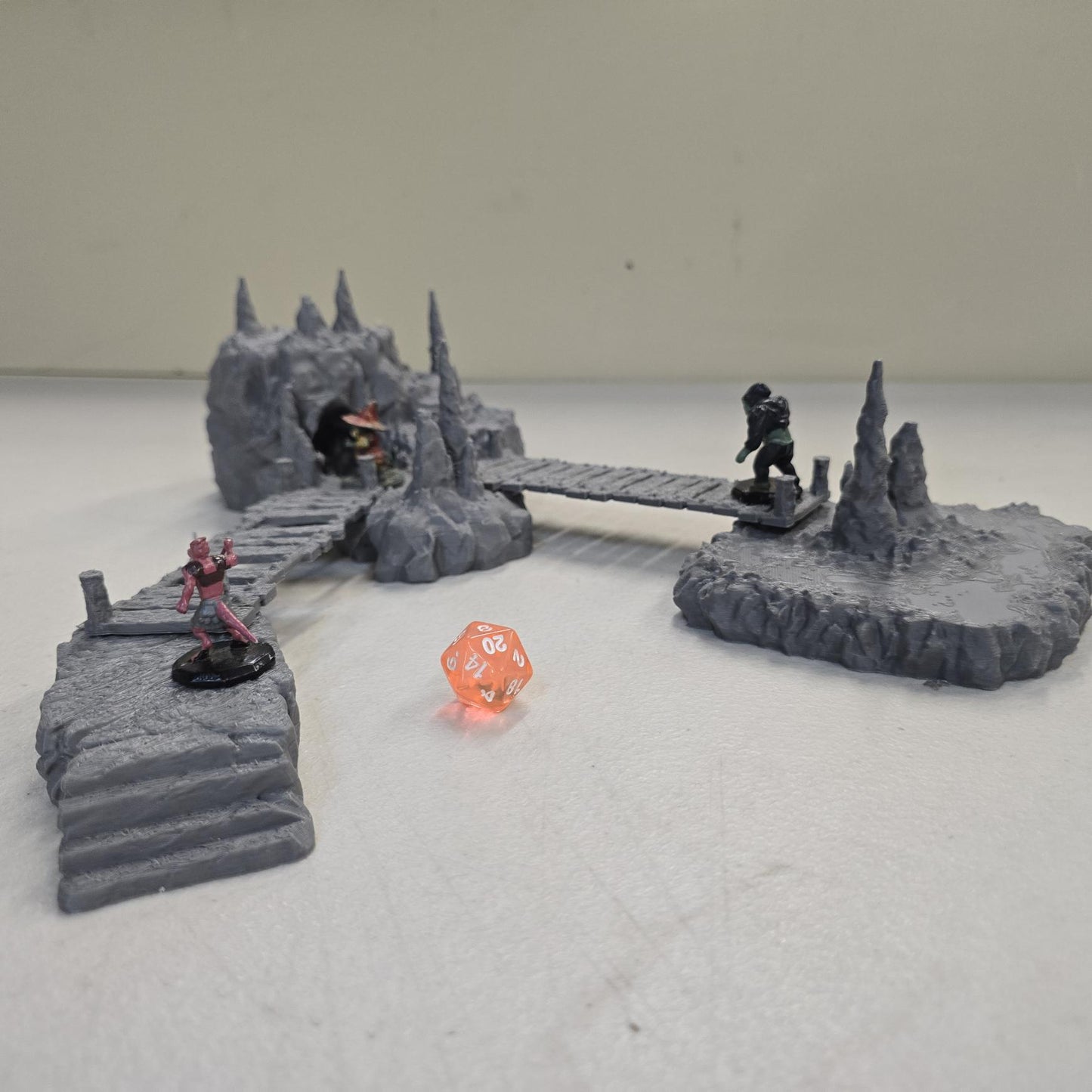 Cavern terrain encounter made from unpainted gray PLA shows a rocky cave entrance surrounded by tall stalagmites and connected by narrow wooden plank bridges. The scene includes a raised stone landing platform and rugged cavern platforms forming a dangerous path across the underground chamber. Scaled for 28 mm tabletop terrain and aligned to a 1-inch grid. The suspended bridges and jagged cavern stone create a tense underground crossing perfect for ambushes and guarded dungeon passages.