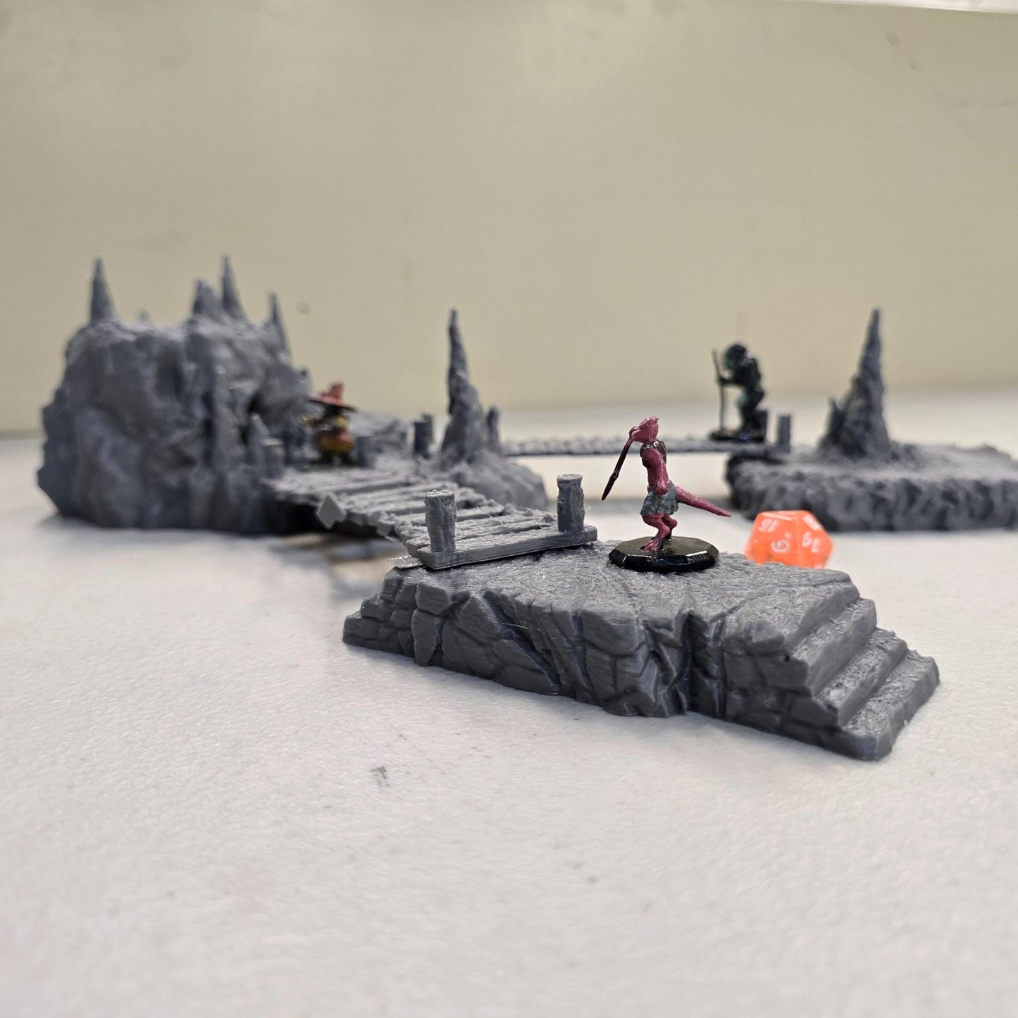 Cavern terrain encounter made from unpainted gray PLA shows a rocky cave entrance surrounded by tall stalagmites and connected by narrow wooden plank bridges. The scene includes a raised stone landing platform and rugged cavern platforms forming a dangerous path across the underground chamber. Scaled for 28 mm tabletop terrain and aligned to a 1-inch grid. The suspended bridges and jagged cavern stone create a tense underground crossing perfect for ambushes and guarded dungeon passages.