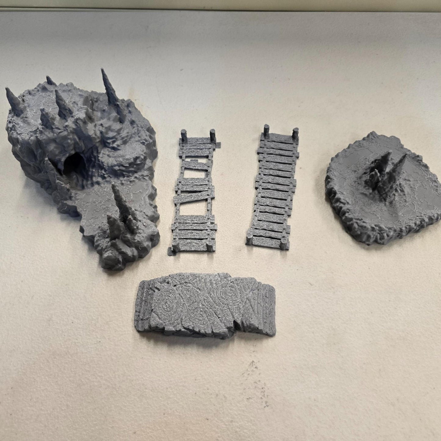 Cavern terrain encounter made from unpainted gray PLA shows a rocky cave entrance surrounded by tall stalagmites and connected by narrow wooden plank bridges. The scene includes a raised stone landing platform and rugged cavern platforms forming a dangerous path across the underground chamber. Scaled for 28 mm tabletop terrain and aligned to a 1-inch grid. The suspended bridges and jagged cavern stone create a tense underground crossing perfect for ambushes and guarded dungeon passages.