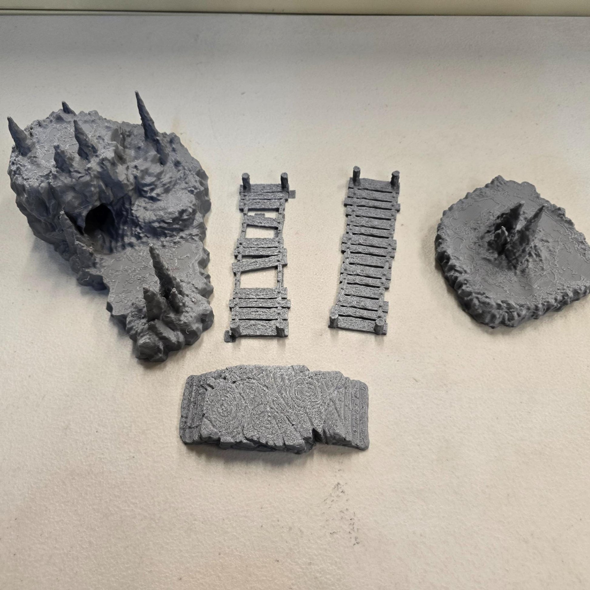 Cavern terrain encounter made from unpainted gray PLA shows a rocky cave entrance surrounded by tall stalagmites and connected by narrow wooden plank bridges. The scene includes a raised stone landing platform and rugged cavern platforms forming a dangerous path across the underground chamber. Scaled for 28 mm tabletop terrain and aligned to a 1-inch grid. The suspended bridges and jagged cavern stone create a tense underground crossing perfect for ambushes and guarded dungeon passages.